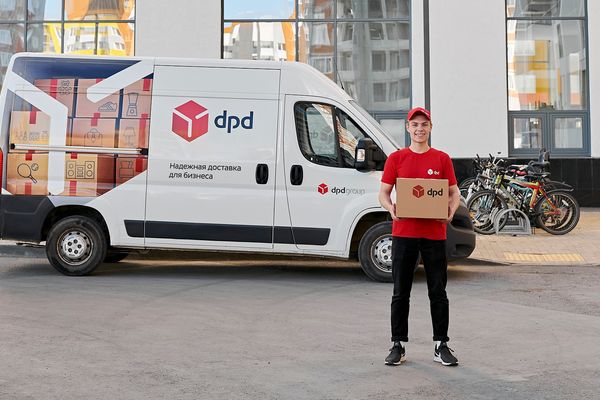 Курьер DPD с посылкой на фоне брендированного фургона компании на улице у жилого комплекса