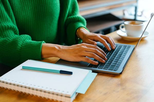Человек в зелёном свитере работает за ноутбуком с блокнотом и чашкой кофе — подготовка аналитического отчета