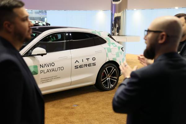 Премиальный кроссовер AITO SERES с брендингом Navio PlayAuto на боковой панели, рядом обсуждают автомобиль двое мужчин в деловой одежде, иллюстрируя запуск мультимедиа с сервисами Сбера