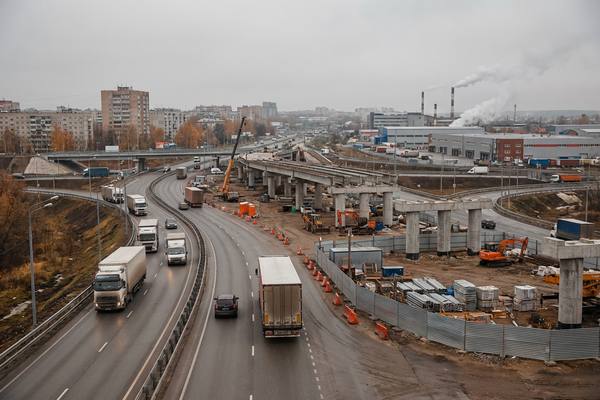 Строительство развязки и эстакады на загруженной городской трассе с грузовиками на фоне промзоны и жилых домов, иллюстрация влияния дефицита региональных бюджетов на дорожную инфраструктуру и логистику