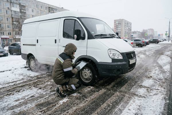 Курьер в зимней одежде проверяет колесо малотоннажного фургона на заснеженной городской дороге