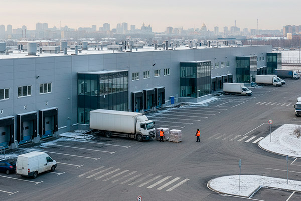 Современный складской комплекс формата light industrial в Санкт-Петербурге зимой, грузовики у погрузочных доков и рабочие в сигнальных жилетах на фоне городской застройки