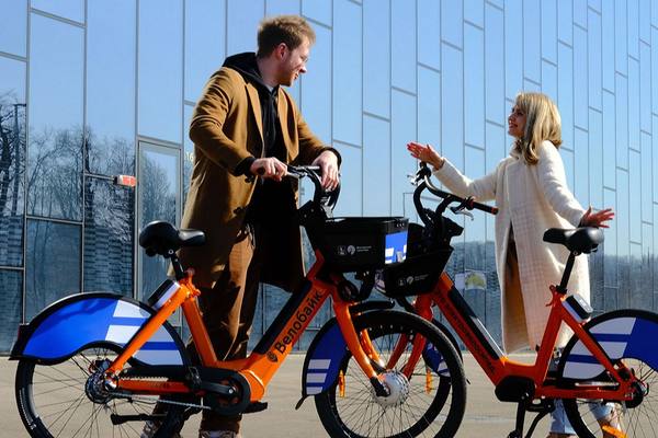 Мужчина и женщина обсуждают городской велопрокат, стоя у велосипедов Ситибайк с логотипом «Велобайк» на фоне современного стеклянного здания
