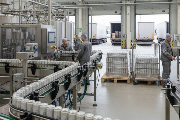 Линия розлива слабоалкогольных коктейлей в алюминиевые банки на российском заводе, паллеты с готовой продукцией и грузовые фуры в зоне отгрузки.