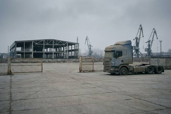 Пустая промышленная площадка с закрытыми воротами, пыльным грузовиком и недостроенным складом на фоне кранов — визуальный образ стагнации и снижения активности в логистике и бизнесе России