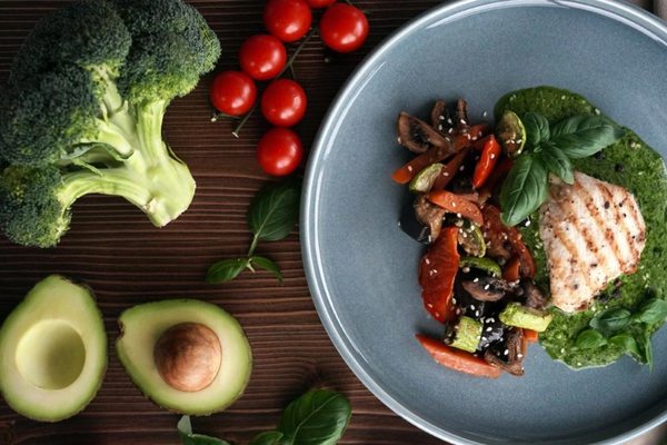 Тарелка с полезным блюдом из запечённого филе и овощей, окружённая свежими продуктами — авокадо, брокколи и томатами; визуальный акцент на здоровом рационе и натуральных ингредиентах.
