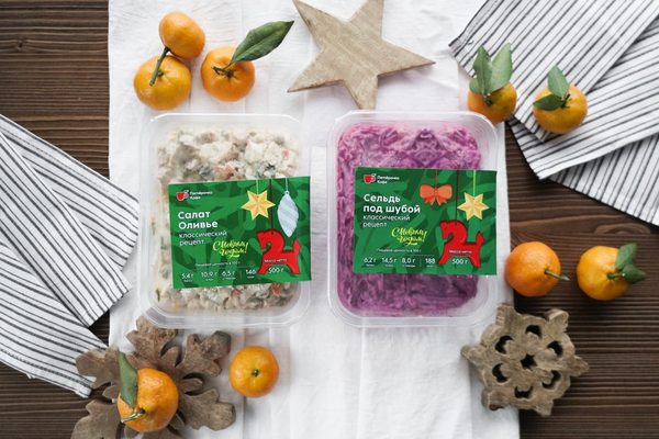 Готовые салаты «Оливье» и «Сельдь под шубой» в праздничной упаковке от «Пятёрочки», размещённые на столе среди новогодних декораций и мандаринов.