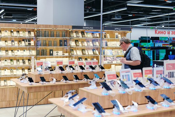 Покупатель рассматривает смартфоны в зале магазина электроники. Витрины с моделями Samsung и Apple, демонстрационные столы с телефонами.