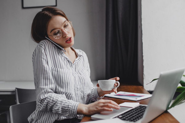 Офисная сотрудница одновременно разговаривает по телефону и работает за ноутбуком в нерабочее время, иллюстрируя размытые цифровые границы между работой и личной жизнью