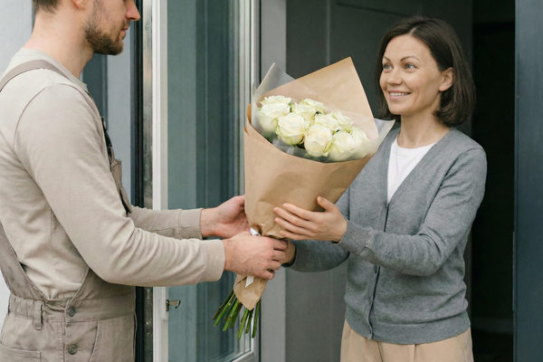Курьер передает букет белых роз клиентке у двери квартиры — доставка цветов как часть последней мили в цветочном бизнесе