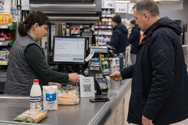 Покупатель оплачивает товары картой на кассе магазина, рядом QR-код СБП и наличные в кассовой зоне