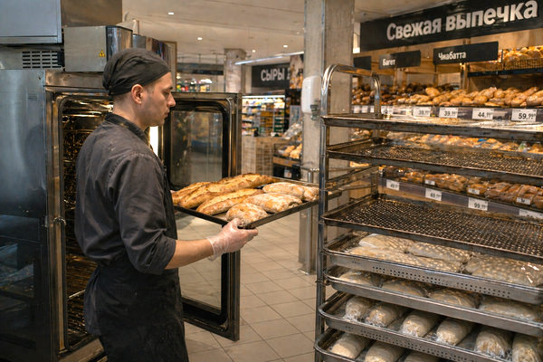 Сотрудник магазинной пекарни в супермаркете вынимает замороженную выпечку после допекания, витрина со свежей выпечкой и хлебобулочными изделиями в ритейле