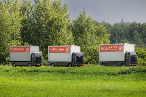 Три автономных грузовика EvoCargo с маркировкой «автономная логистика» движутся вдоль зеленого поля на фоне леса в Подмосковье